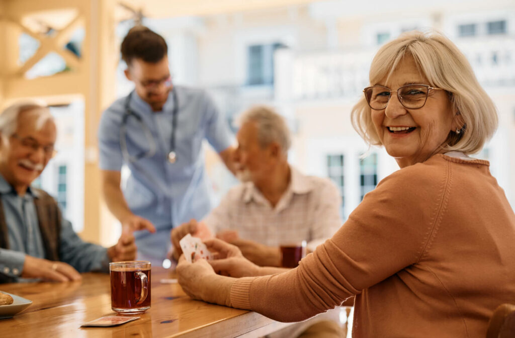 A group of older adults at a senior living community sit around a table laughing and playing cards togethe