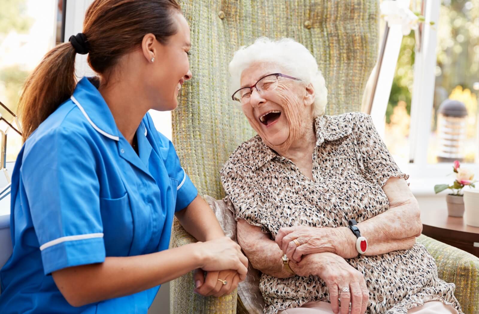 An older adult woman in a senior living facility sitting on a chair, smiling, and having a conversation with a nurse.