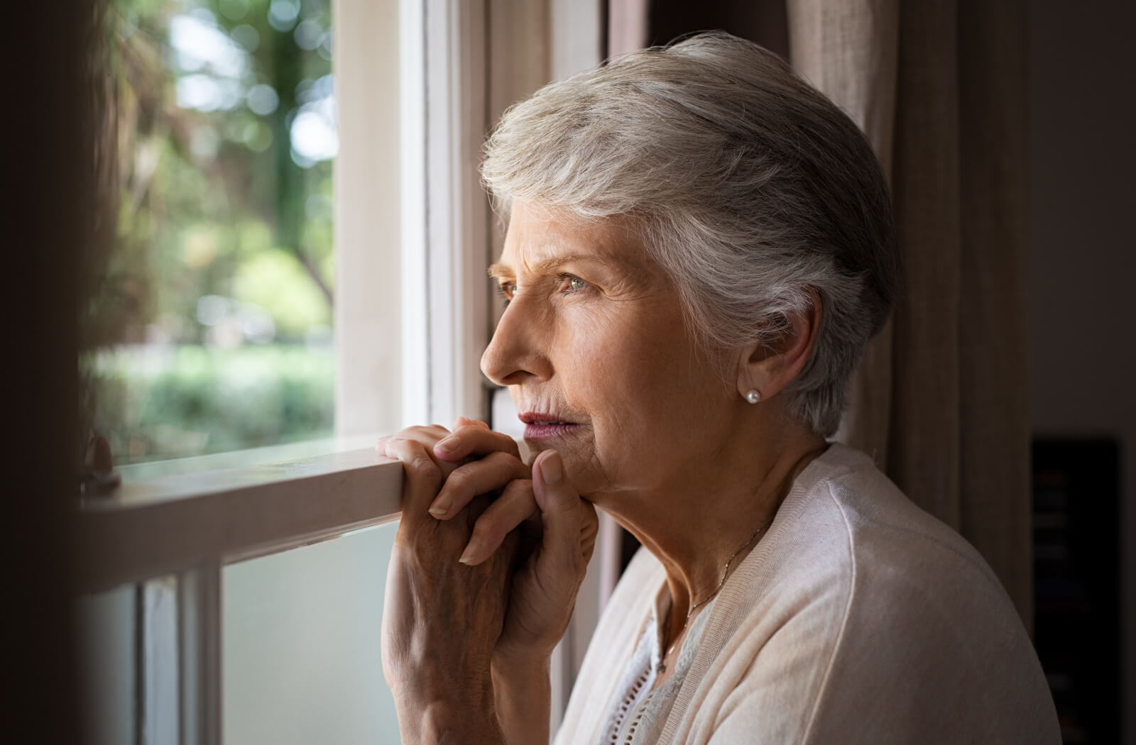 An older adult woman holding onto a window sill and looking out of the window with a serious expression