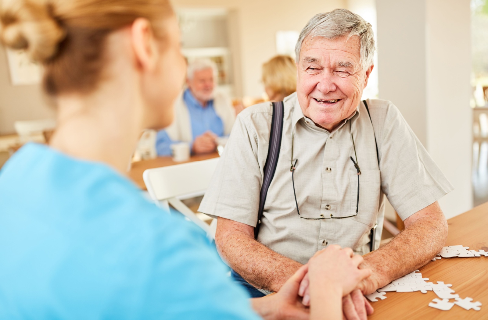 A staff member in memory care and a smiling parent with dementia holding hands while seated at a table with puzzle pieces.
