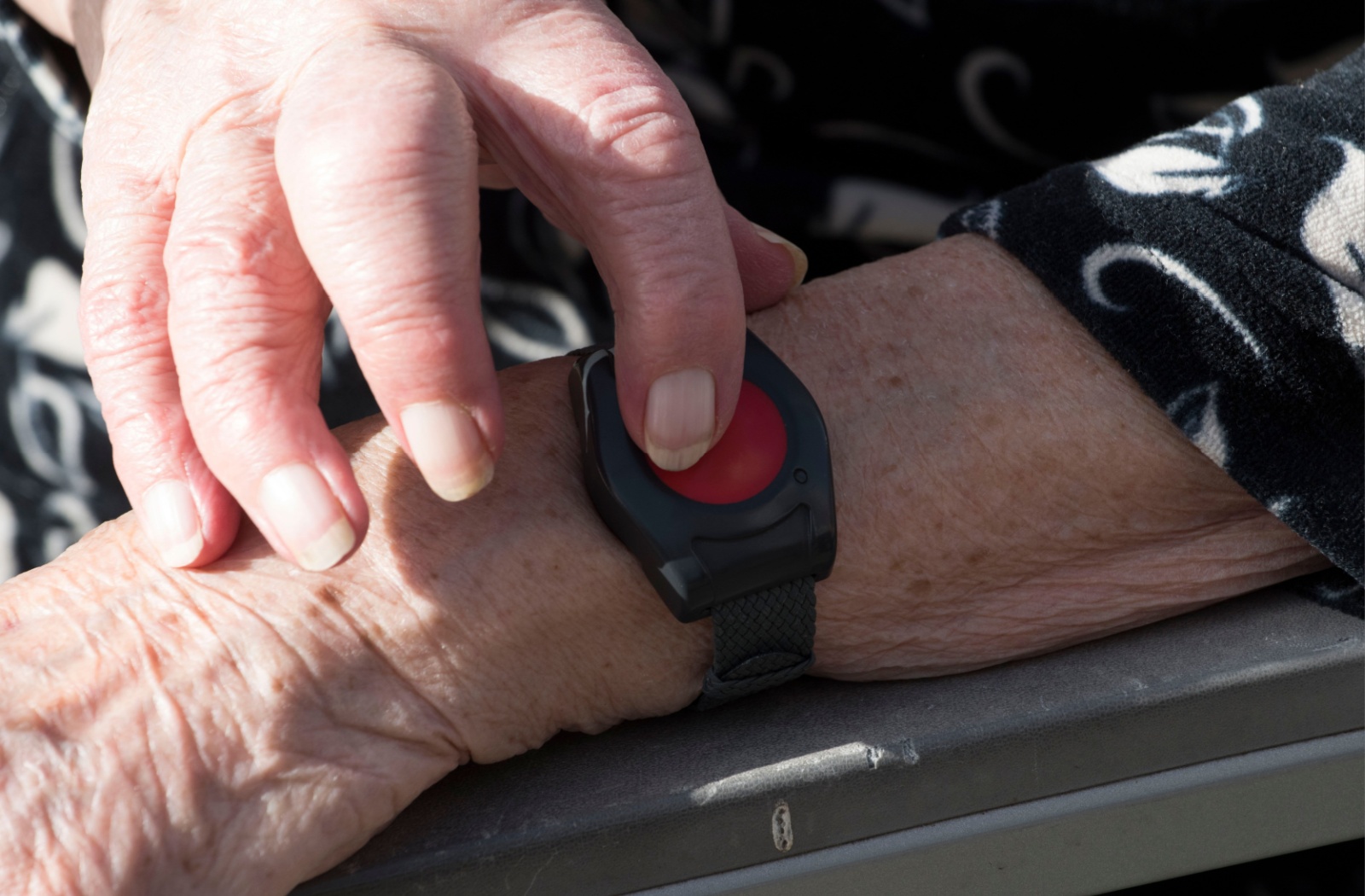 A close up of an older adult's left hand with a medical alert bracelet and right index finger pressing on a red button.