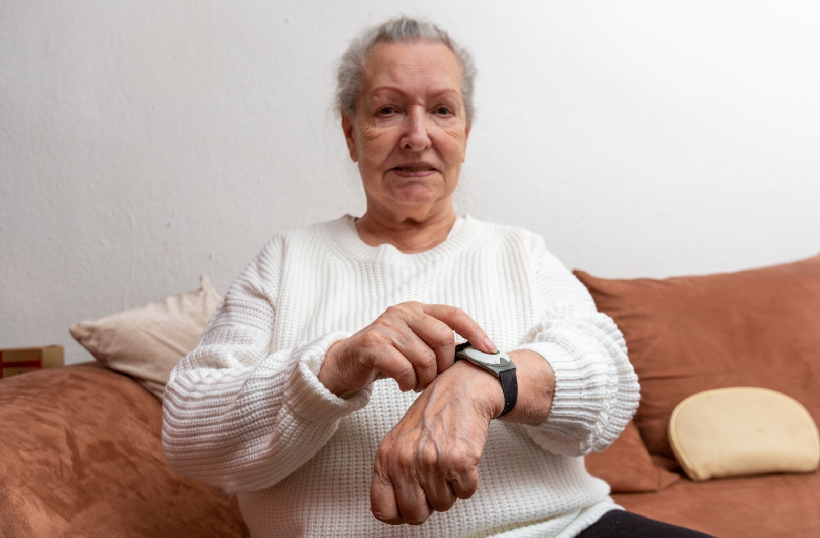 An older adult sitting on a couch pointing to her medical alert bracelet on her left hand with her right index finger.
