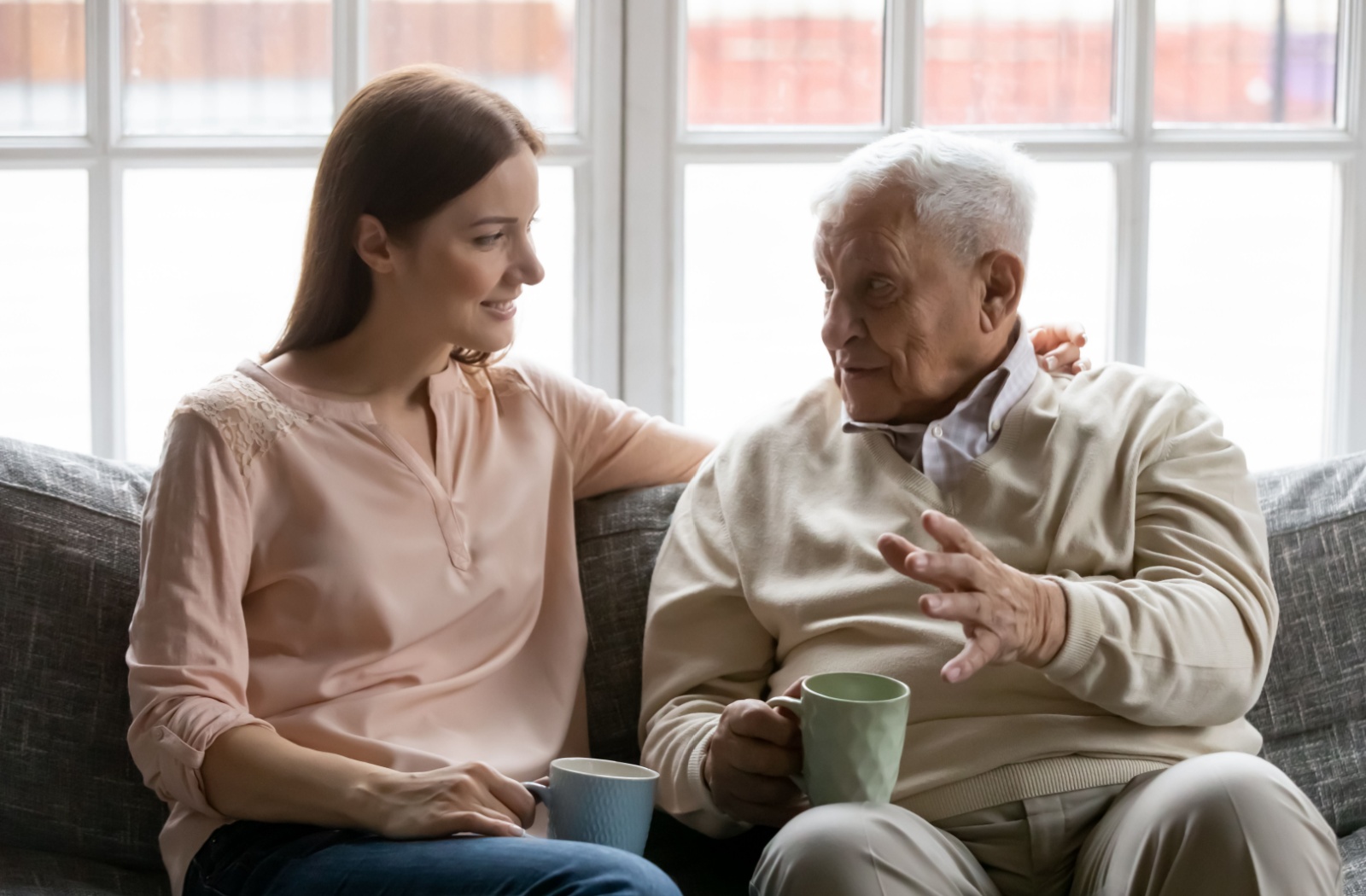 An adult child and her father hold mugs of tea and sit on the couch during a conversation while she has her left arm over his shoulder.