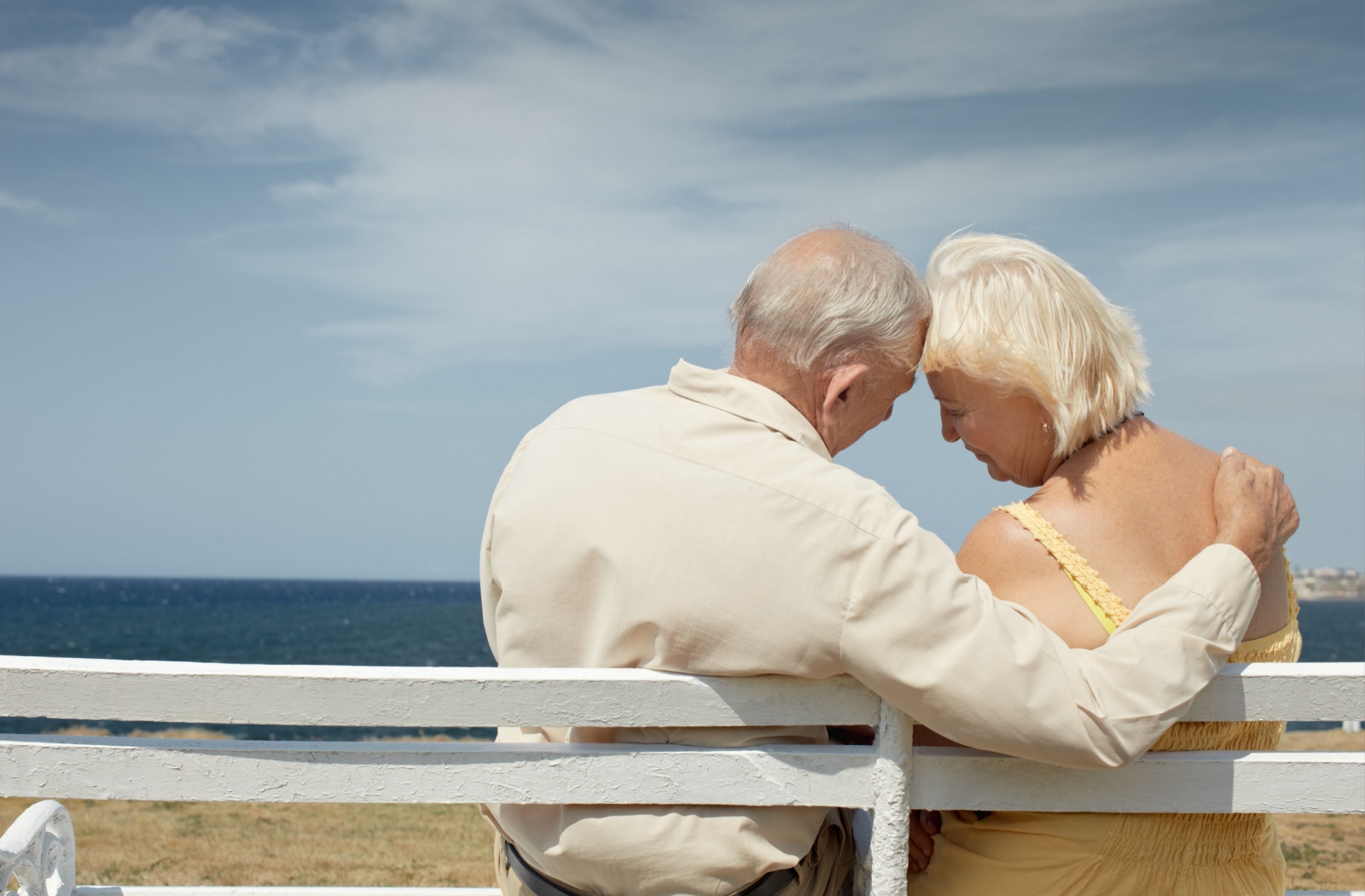 A senior couple enjoys an afternoon sitting on a bench by the ocean.