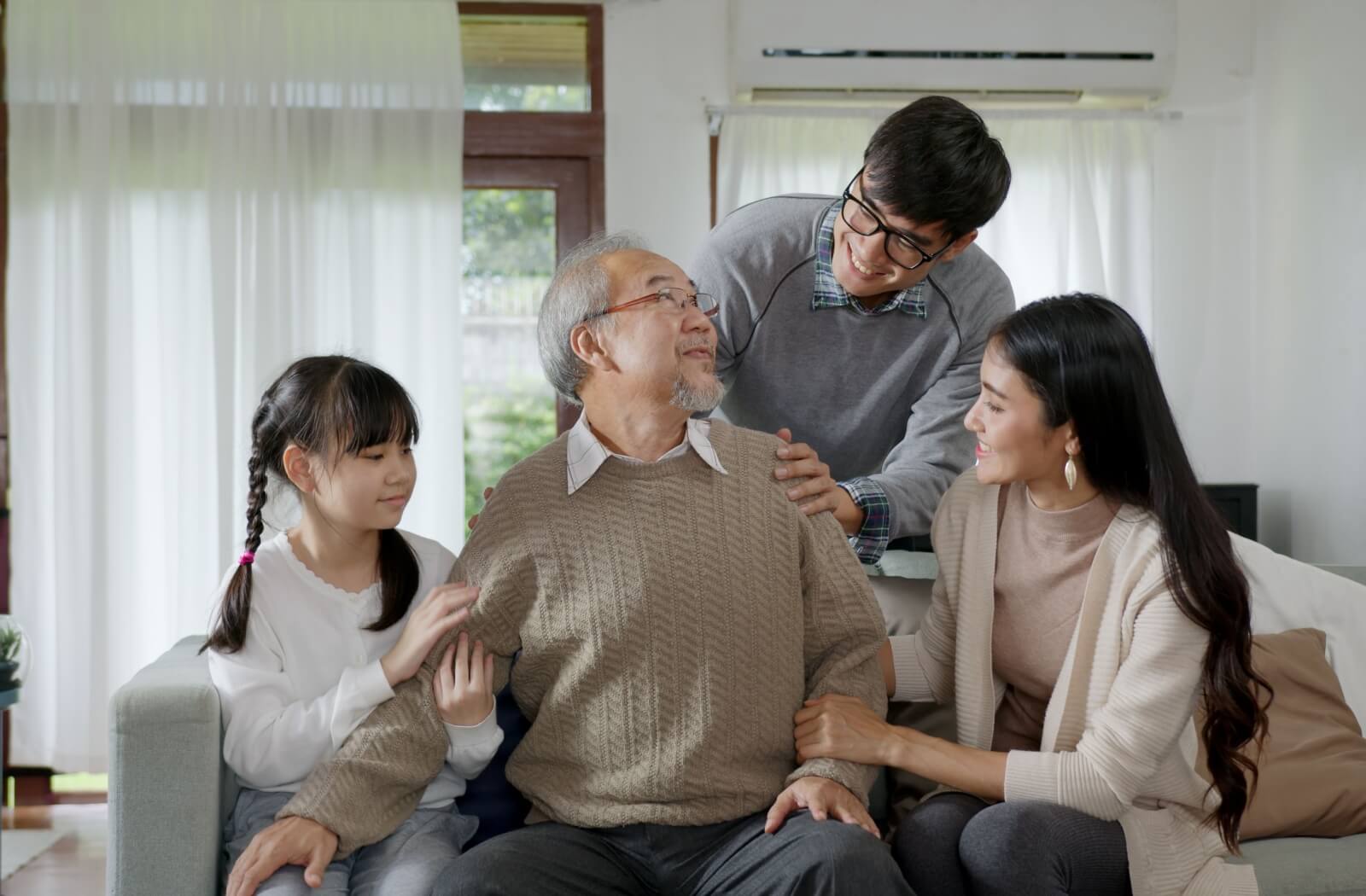 A multi-generational family comforts an elderly person sitting on a couch in a bright, cozy setting

