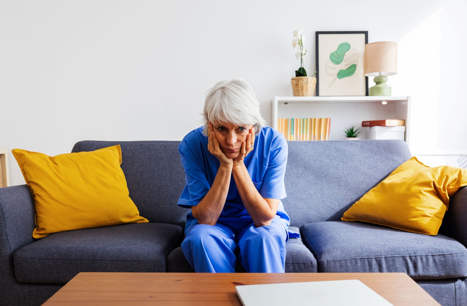 An exhausted caregiver in blue clothes sits in the middle of a couch, resting their head on both hands