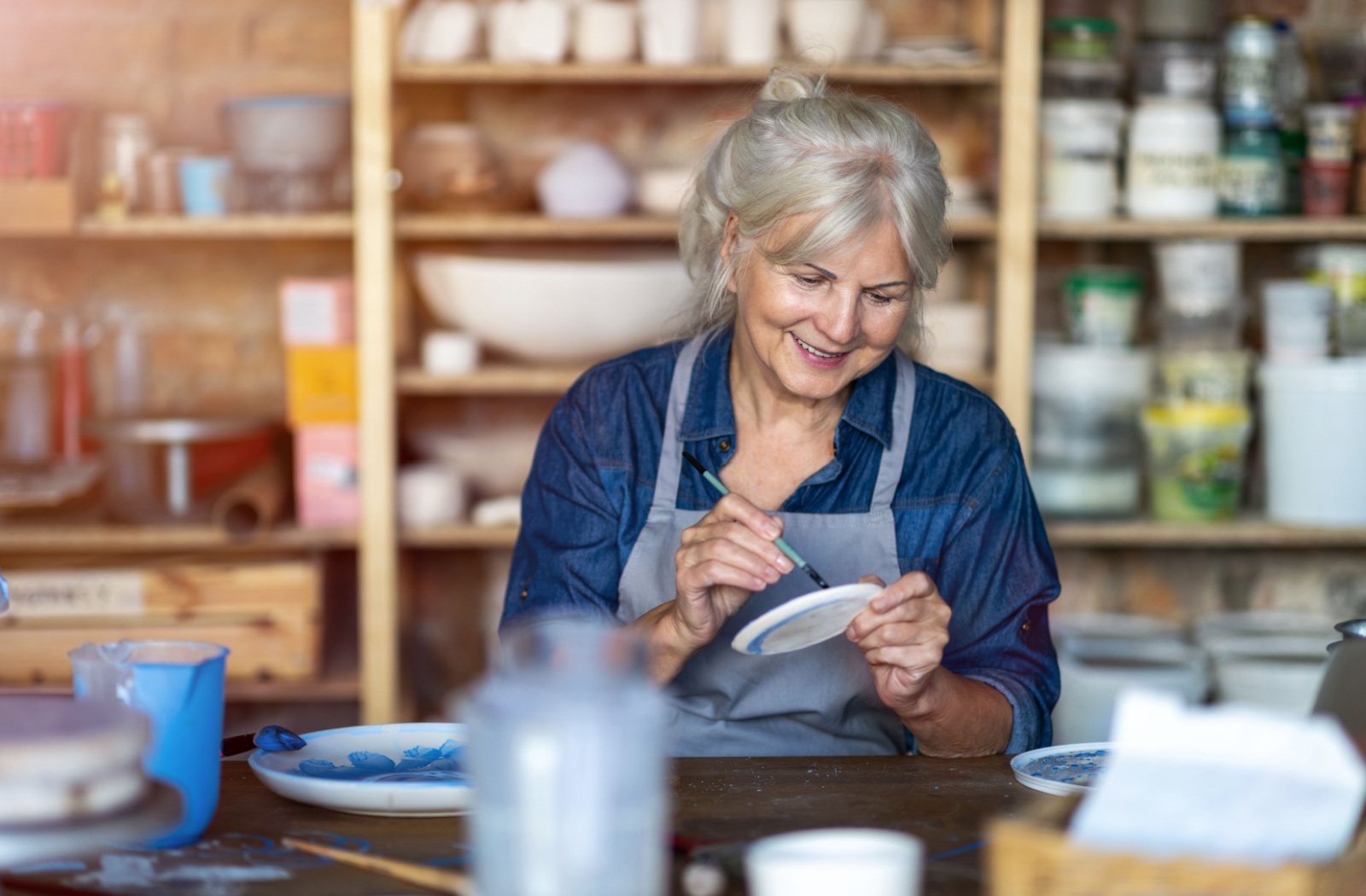 Senior painting a ceramic plate blue while smiling in an art studio surrounded by pottery and supplies