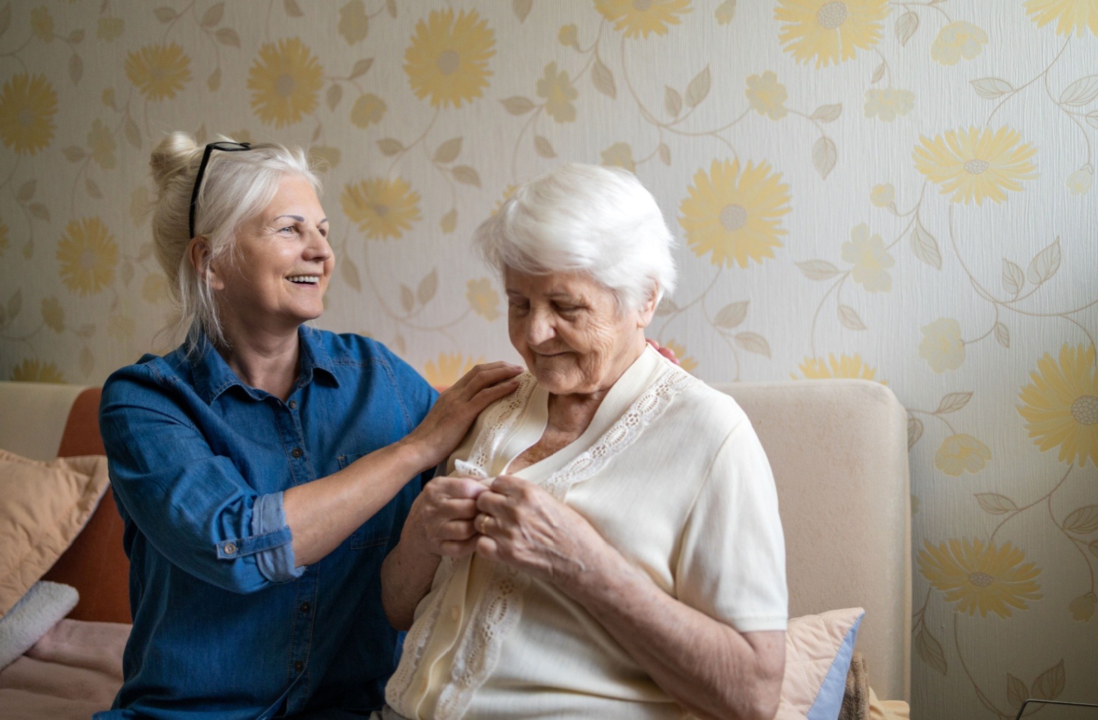 An adult child comforts their parent with dementia as they button up their blouse.